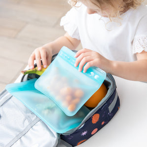 A child packs snacks into an eco-friendly lunchbox using Bumkins Silicone Flat Reusable Bags with a child-friendly zip seal.