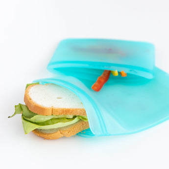 A sandwich with lettuce in a partially open Bumkins Silicone Flat Reusable Bag, Blue, next to carrot sticks in an eco-friendly setup.