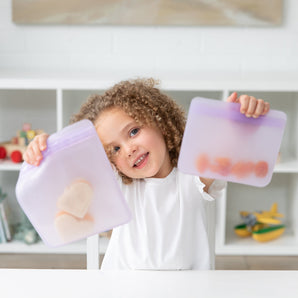 Child happily holds two Bumkins Lavender Silicone Flat Reusable Bags full of snacks, sitting at a white table with shelves behind.