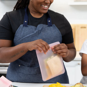 A person in blue apron places a sandwich into a Bumkins Silicone Flat Reusable Bag with a child-friendly zip seal.