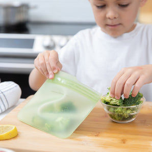 A child puts broccoli in a Bumkins Silicone Flat Reusable Bag, Sage, on the kitchen counter.