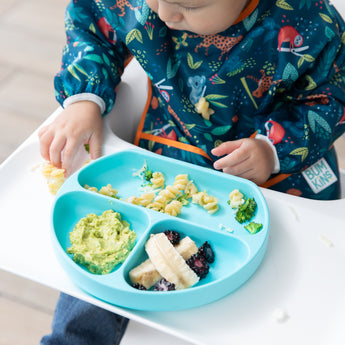 A child in a colorful bib enjoys pasta and broccoli from the blue Bumkins Silicone Grip Dish with a secure suction base.