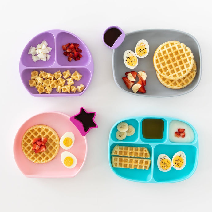 Four colorful divided plates with waffles, fruit, eggs, and dipping sauces arranged aesthetically. The plates are purple, gray, pink, and blue, each containing a variety of breakfast items like strawberries, bananas, and syrup.