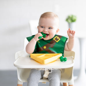 Baby in a high chair, wearing a green-brown bib, smiling and holding a toy, with the Bumkins Zelda™ Triforce Silicone Grip Dish.