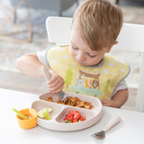 A toddler in a yellow bib enjoys food from a Bumkins Silicone Grip Dish: Sand, filled with pancakes, strawberries, and pears.