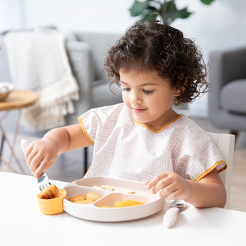 A young child eagerly eats from a Bumkins Silicone Grip Dish: Sand in a cozy living room.