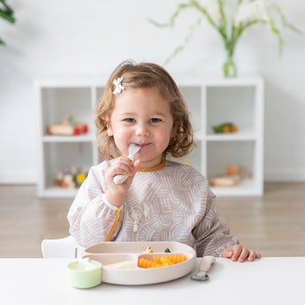 A toddler with a flower clip enjoys a meal from her Bumkins Silicone Grip Dish: Sand, securely attached to the table by its suction base.