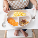 Child in high chair with Bumkins Silicone Grip Dish: Sand, holding scrambled eggs, orange slices & star pancake. Spoon and fork ready.