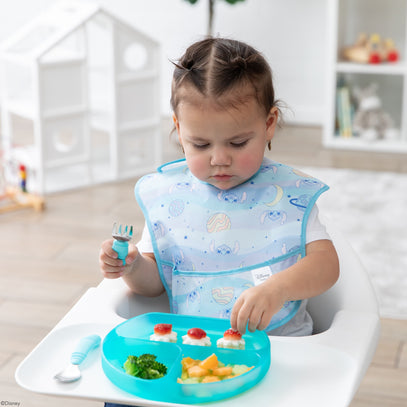 A toddler with pigtails sits in a high chair, holding a fork with a blue handle. The child wears a bib with a cute pattern and focuses on a turquoise plate with broccoli, cheese, and snacks topped with sauce. A bright, cozy room is in the background.