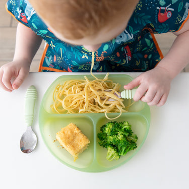 A child with a colorful bib is enjoying spaghetti from the Bumkins Silicone Grip Dish: Green Jelly, designed for baby and toddler portions. Its strong suction base secures broccoli and cornbread while the child eats with a fork, and a spoon rests beside the plate.