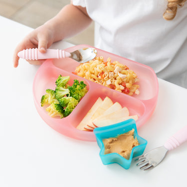 A child enjoys a meal from a pink Silicone Grip Dish: Pink Jelly by Bumkins, with divided sections for macaroni, broccoli, and apple slices. A nearby blue star-shaped dish holds peanut butter. The LFGB silicone plates suction base keeps it steady on the white table as the child sits with a spoon in hand.