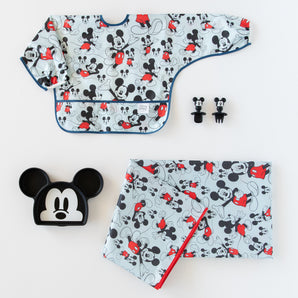 The Bumkins Disney Little Ones Gift Bundle: Mickey Mouse Classic includes a themed sleeved bib, two pacifiers, a black Mickey-shaped plate, and a matching bag with a red zipper. Featuring gray, black, red, and white hues with cartoon designs. Ideal for young fans!.