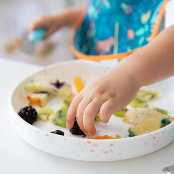 A child reaches for kiwi & blackberry on a Bumkins Vanilla Sprinkle Silicone Grip Plate, secured with suction, while wearing a colorful bib.