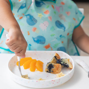 A childs hand uses a fork to pick food from a Bumkins Silicone Grip Plate: Vanilla Sprinkle, wearing a sea creature bib.