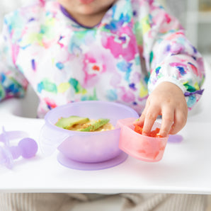 A child in a colorful shirt reaches for food from a Bumkins Silicone Little Dipper Combo on a white surface, using the Magenta Jelly pack.