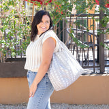 A person with shoulder-length dark hair stands smiling outdoors, wearing a light-colored, sleeveless knit top and jeans. Theyre carrying a large gray Packable Tote Bag: Lunar Phase by Bumkins. Greenery and a fence appear in the background.