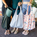 Three individuals, dressed in casual athletic wear and sandals, stand close on a brick walkway each holding Bumkins Packable Tote Bags. The designs they showcase are a dark pattern, one with light circles, and the Lunar Phase pink print. A yoga mat peeks from each bag.