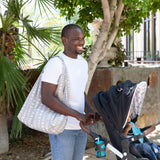 A man in a white shirt pushes a stroller with Bumkins Packable Tote Bag: Lunar Phase on his shoulder in a sunny outdoor setting with trees.