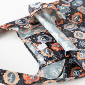 Close-up of the Bumkins Packable Tote Bag: Ghostly Gallery, showcasing a Halloween pattern with skulls, pumpkins, and decorative frames in orange, black, and gray. The bag is slightly open, revealing a gray waterproof interior.