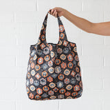 A person holds a Bumkins Packable Tote Bag: Ghostly Gallery, featuring a colorful animal pattern on black fabric with orange and blue accents. The waterproof tote is displayed against a textured white brick wall.