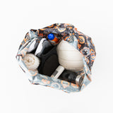 Top view of a Bumkins Packable Tote Bag: Ghostly Gallery, showcasing waterproof fabric, filled with a white volleyball, rolled towel, black and white sports shoes, water bottle, and sunscreen. The multicolored sports-themed design adds flair reminiscent of Disneys Nightmare Before Christmas.