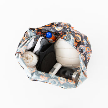 Top view of a Bumkins Packable Tote Bag: Ghostly Gallery, showcasing waterproof fabric, filled with a white volleyball, rolled towel, black and white sports shoes, water bottle, and sunscreen. The multicolored sports-themed design adds flair reminiscent of Disneys Nightmare Before Christmas.