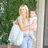 A woman smiles, carrying a child and a Bumkins Packable Tote Bag: Aloha Stitch near lush foliage.