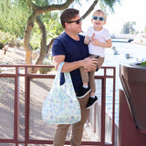 A man holds a smiling child, both wearing sunglasses, with a Bumkins Packable Tote Bag: Aloha Stitch on his arm. 