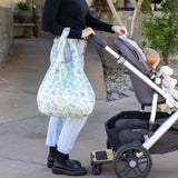 A person strolls outdoors with a baby in a stroller, carrying a Bumkins Packable Tote Bag: Aloha Stitch over their arm.