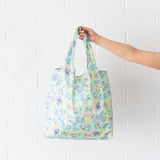 A hand holds a Bumkins Packable Tote Bag: Aloha Stitch against a white brick wall.