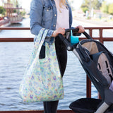 Person in denim jacket by the canal, pushing a stroller and holding a Bumkins Packable Tote Bag: Aloha Stitch.