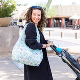 A woman with curly hair in a black sweater pushes a stroller, carrying the Bumkins Packable Tote Bag: Aloha Stitch and wearing sunglasses.