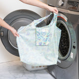 A person holds the Bumkins Packable Tote Bag: Aloha Stitch in front of an open washing machine.