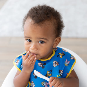 A toddler in a blue Bumkins SuperBib® 3 Pack: Mickey & Friends MVP eats with a spoon, wearing a curious expression.