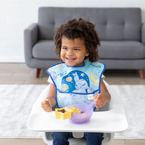 A curly-haired child smiles in a high chair, wearing a Bumkins SuperBib® 3 Pack: Stitch Star-ring Up Trouble and eating from a purple bowl.