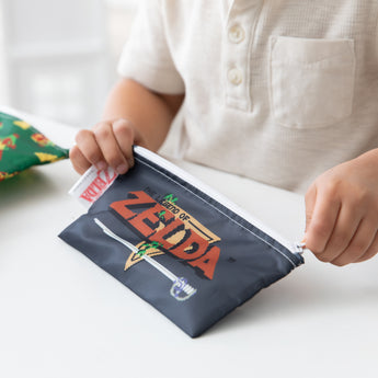 Child in white shirt holds a Bumkins Reusable Snack Bag featuring The Legend of Zelda design, made from waterproof fabric.