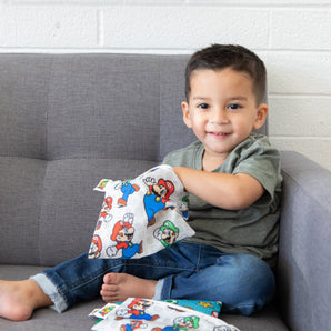 A smiling child sits on a gray couch, clutching a pouch with colorful animated characters like on Bumkins Super Mario™ & Luigi bag.