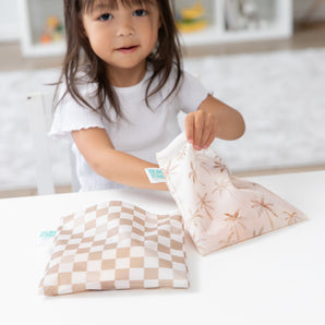 A child places Bumkins Reusable Snack Bag, Large 2 Pack: Palm Check on a table.