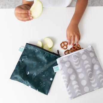 A child reaches for pretzels from a white snack bag featuring a moon phase design and apple slices over Bumkins Desert Night large reusable snack bag, crafted from waterproof fabric. Both bags on the white table blend style and practicality with the convenience of easy machine washing.