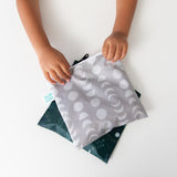 A child holds a Bumkins Large Reusable Snack Bag, Desert Night design, above a flat dark green pouch on a white surface.