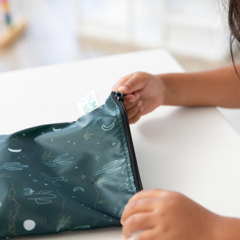 A child eagerly unzips a Bumkins Desert Night reusable snack bag, large size, showcasing charming white cacti, moon, and star illustrations on waterproof dark green fabric. Conveniently machine washable, this delightful pouch rests on a white table.