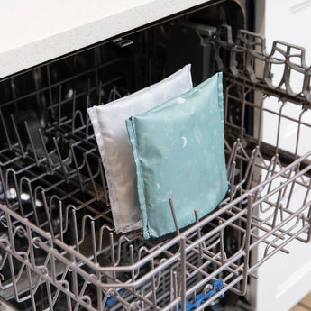 Two Bumkins Reusable Snack Bag, Large 2 Pack: Desert Night bags, made from waterproof fabric with light gray and teal patterns, are placed on the top rack of an open dishwasher, highlighting their easy machine wash care while the dishwasher remains otherwise empty.