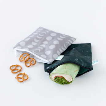 The two large reusable snack bags from Bumkins Desert Night pack, one gray with a moon pattern and one black with a celestial design, are partially open on the white surface. The waterproof black bag holds a tortilla wrap with greens and deli meat, while five pretzels are scattered nearby. They’re machine washable after use.