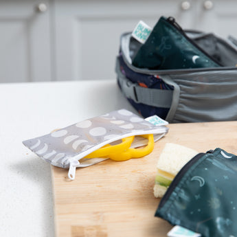 Bumkins Desert Night reusable snack bags sit on the kitchen counter, one open with a banana inside, next to a packed sandwich.