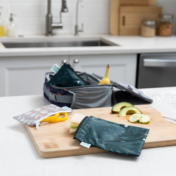 Bumkins Reusable Snack Bag, Large 2 Pack: Desert Night, with green and grey patterns rests on a kitchen counter; one contains a sandwich, another sliced cucumbers, and the third a banana. They are made from waterproof fabric and are machine washable. A sink and cutting board can be seen in the background.