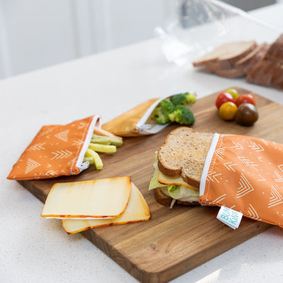 Reusable orange sandwich bags with white arrow patterns are on a wooden board. One bag holds a sandwich, another has broccoli, and the third has sliced cheese. Cherry tomatoes and sliced bread are in the background.