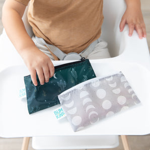 A child in a high chair reaches for Bumkins Reusable Snack Bag, Small 2 Pack: Desert Night on the tray.