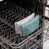 Two Bumkins Desert Night reusable snack bags sit on a dishwasher top rack beside two packs of sanitary pads.