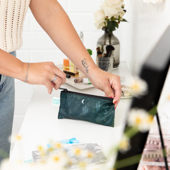 Person arranging a Bumkins Desert Night Reusable Snack Bag on a makeup table, surrounded by flowers and travel organizers.