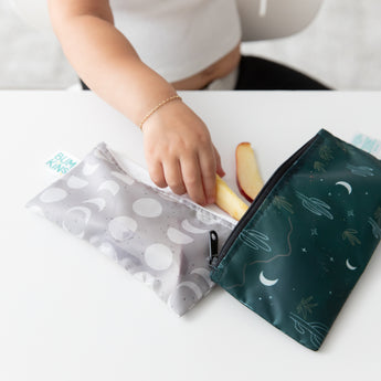 A child in a white top and gold bracelet reaches for apple slices from Bumkins Desert Night reusable snack bag with green patterns, while a gray bag with white circles is nearby, ready for storage and travel. These handy organizers provide convenience on the go.